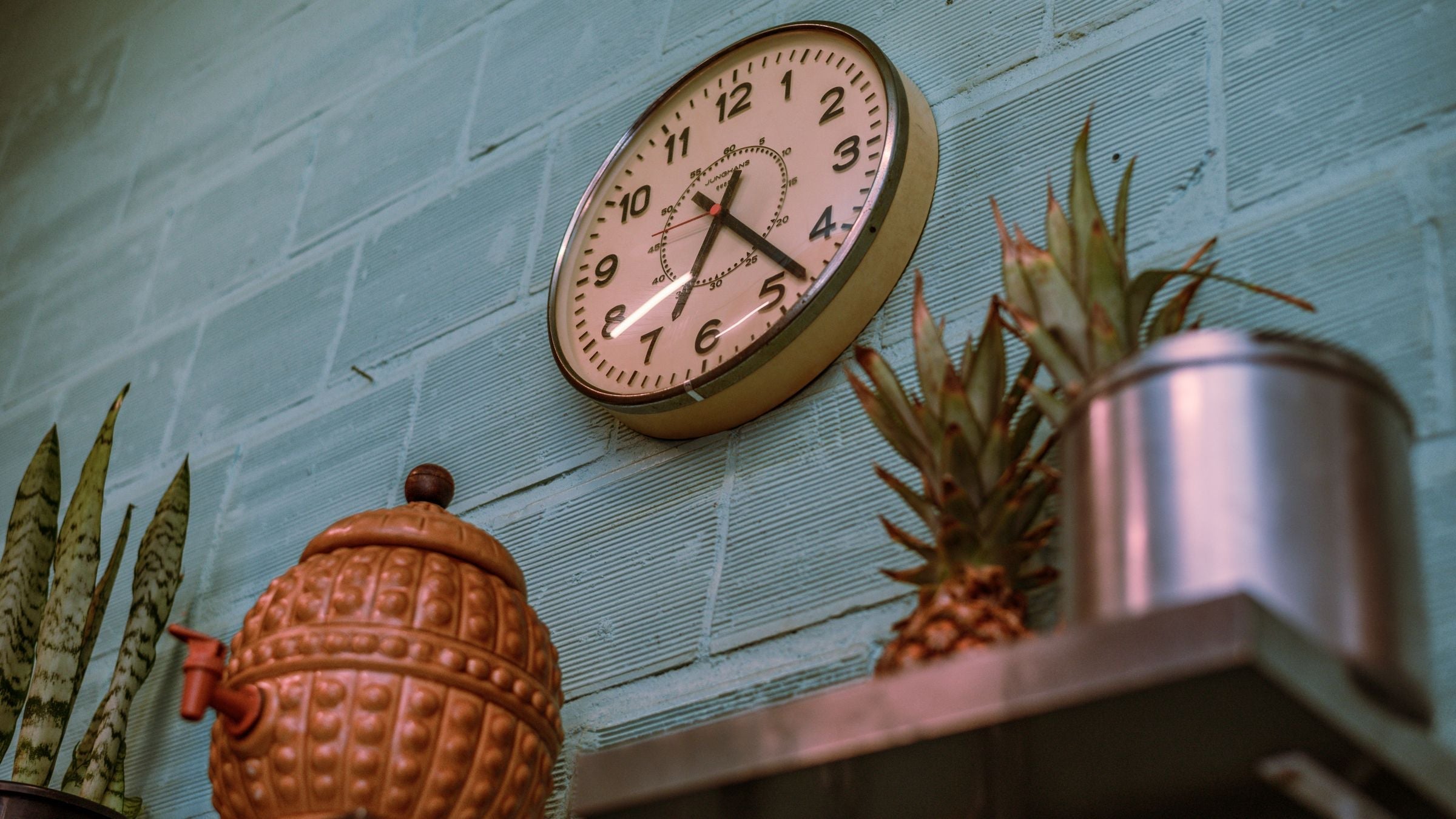 Horloge murale vintage au cadran crème sur un mur de briques céladon, entre plantes et pichet en terre cuite