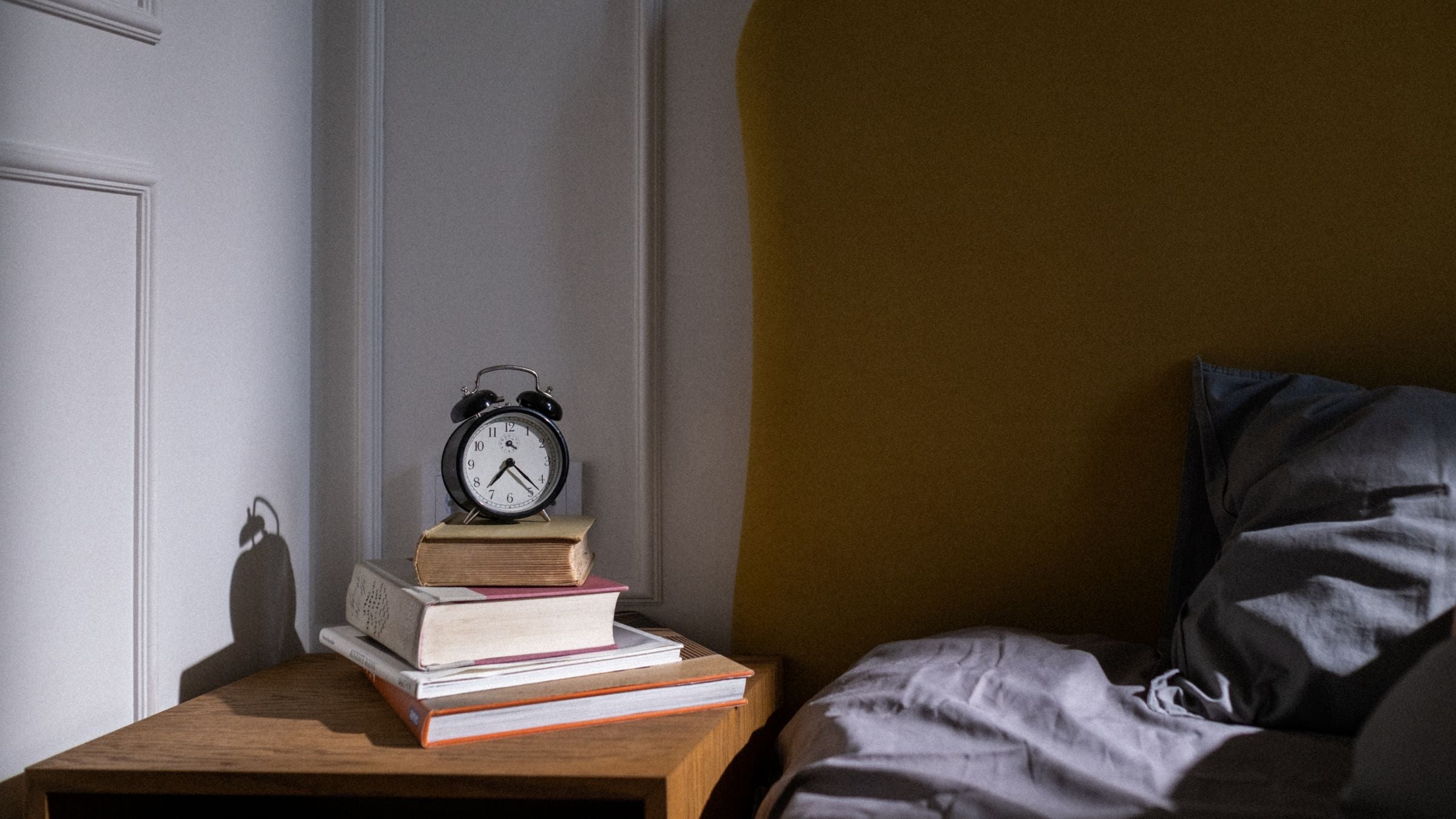 Réveil à cloches posé sur une pile de livres sur la table de nuit, à côté d'un lit au petit matin