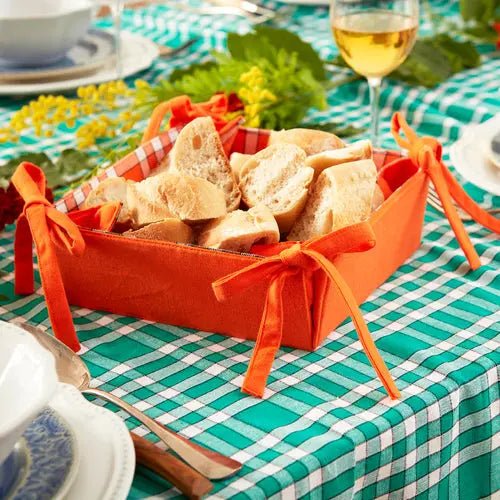 Corbeille à pain en tissu orange sur table à carreaux, avec des morceaux de pain et décorée de rubans, idéale pour présentation élégante.