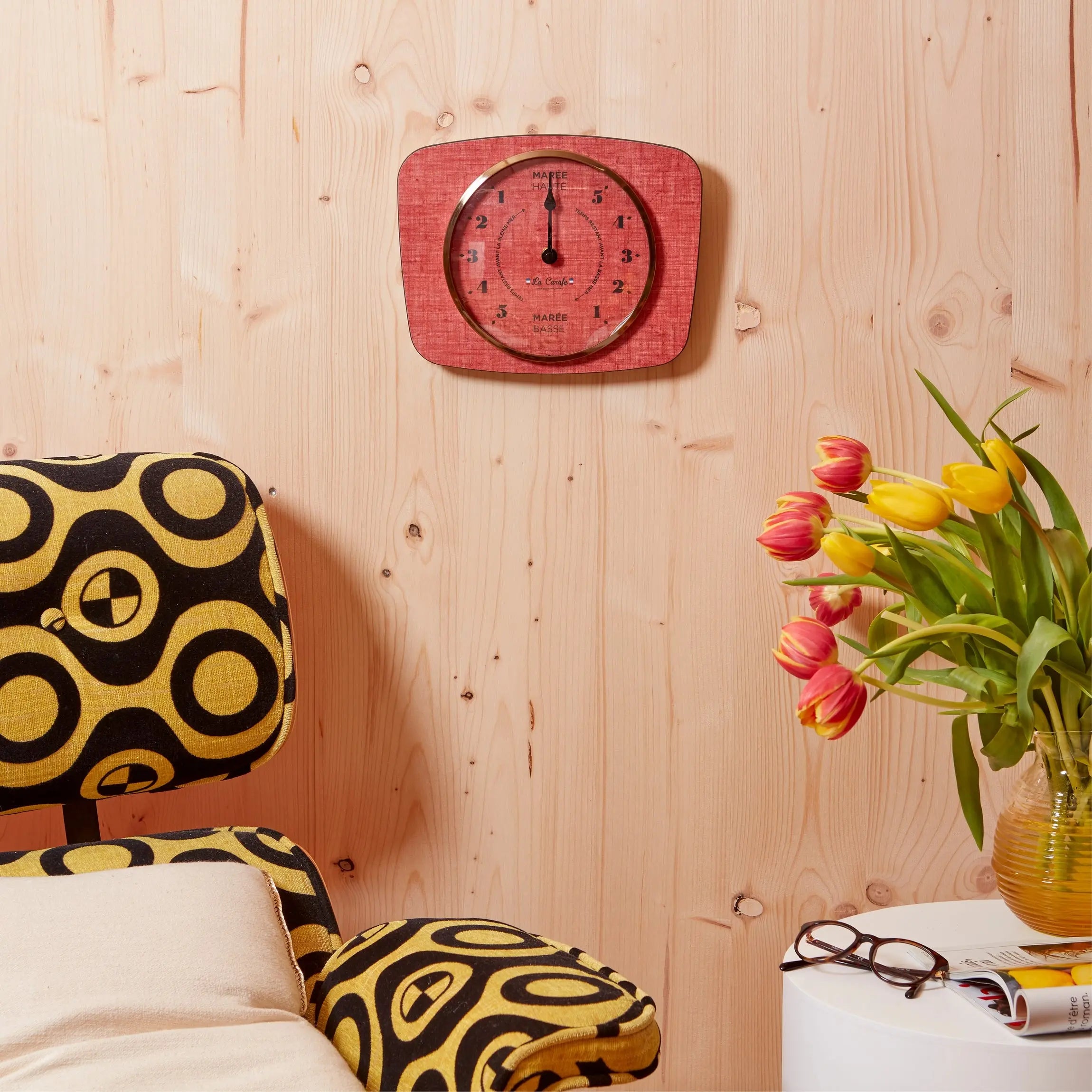 Horloge des Marées murale rouge avec affichage des cycles de marée, déco élégante sur un mur en bois avec fauteuil et fleurs colorées.