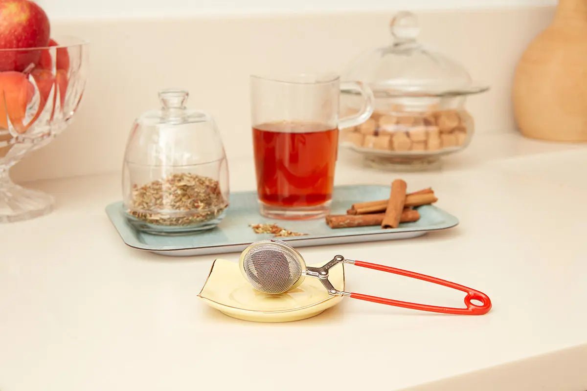 Pince à thé rouge sur un plateau avec une tasse de thé et des épices, accessoire pratique pour infusion élégante.