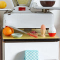 Râpe acier inoxydable rétro posée sur plan vintage avec orange, œuf, minuterie et décoration maison années 50-70.